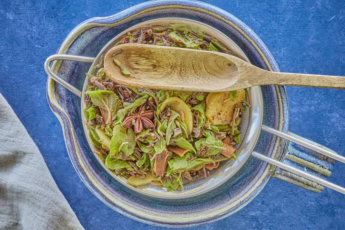a mixture of steeped leaves and spices in a colander over a large blue bowl, with a wooden spoon on top of the spice mixture, and a kitchen towel in the background