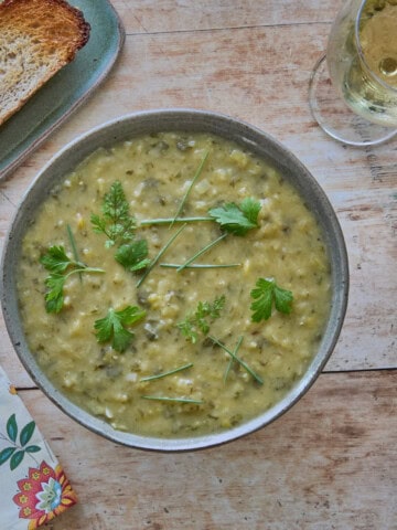 a square photo of a bowl of soup with fresh herb garnishes, with a napkin, toasted piece of bread on a small plate, glass of white wine, and spoon on the table around the bowl