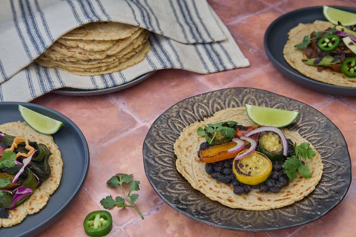 three plates of tortillas with various veggie toppings, garnishes in front, and a plate of kitchen towel-wrapped tortillas in the background