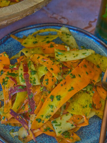 a close-up shot of carrot ribbons in chermoula sauce on a plate with a fork, a large wooden bowl of carrots, a napkin, and a glass of mint tea