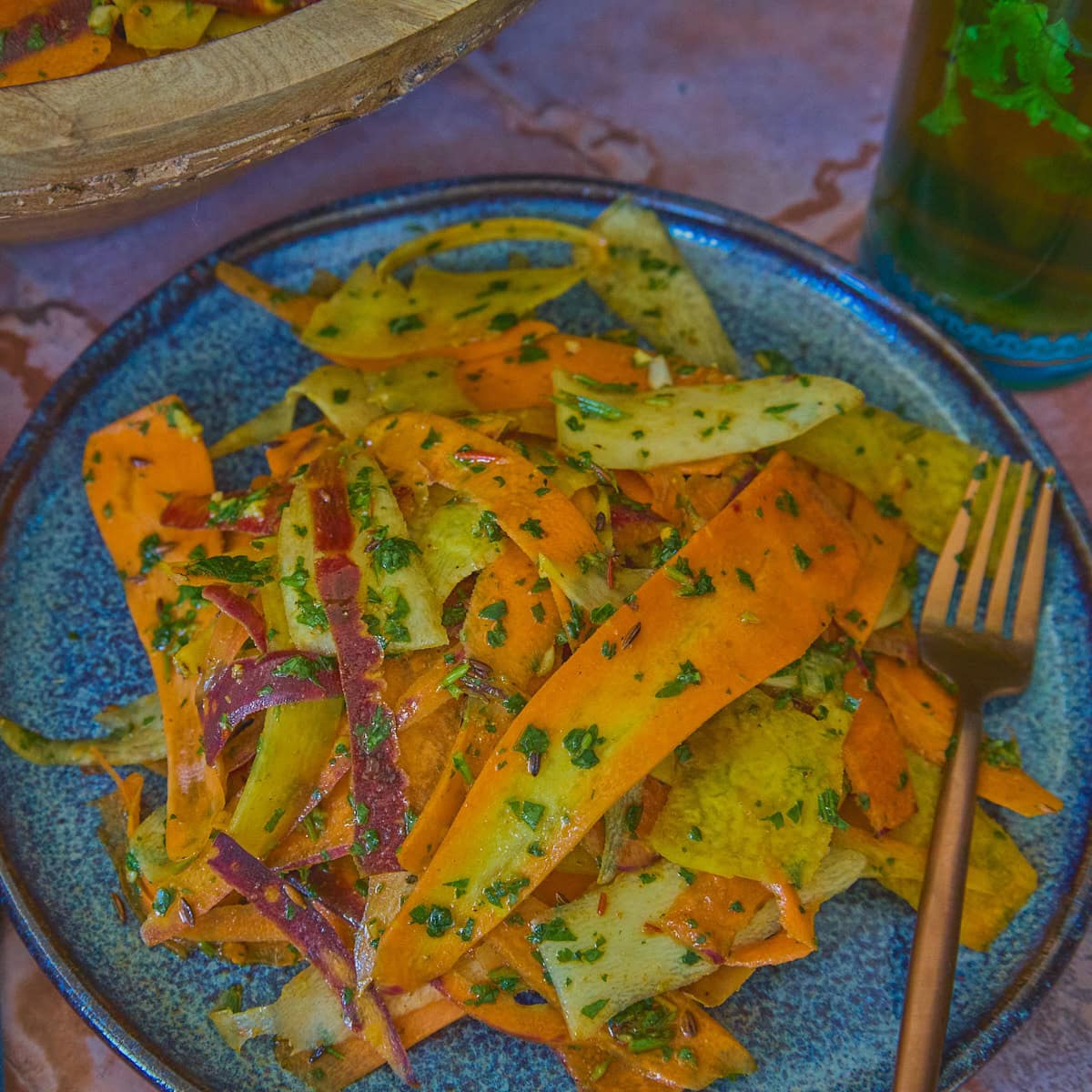 a close-up shot of carrot ribbons in chermoula sauce on a plate with a fork, a large wooden bowl of carrots, a napkin, and a glass of mint tea