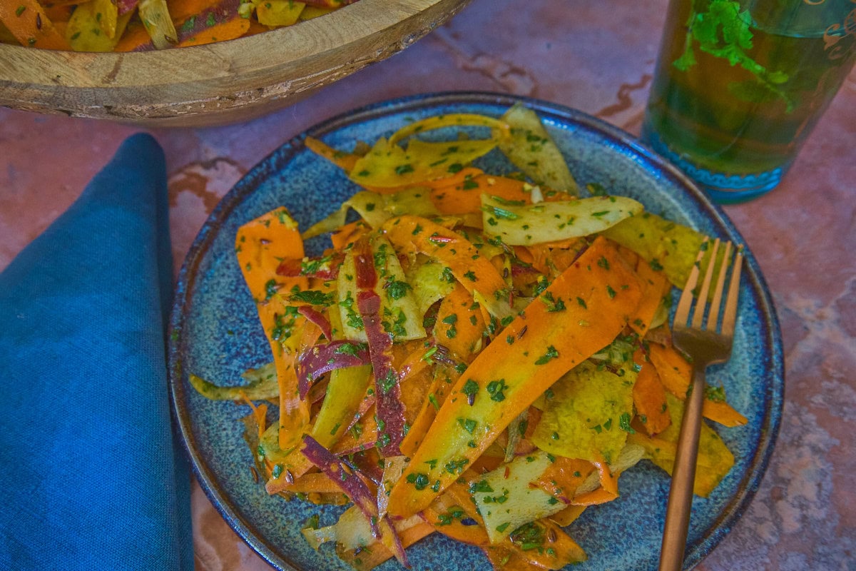 carrots with chermoula sauce on a plate with a fork, a large wooden bowl of carrots, a glass of mint tea, and a napkin