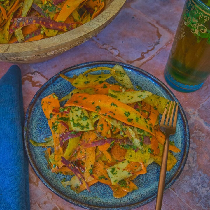 carrot ribbons in chermoula sauce on a plate with a fork, a large wooden bowl of carrots, a napkin, and a glass of mint tea