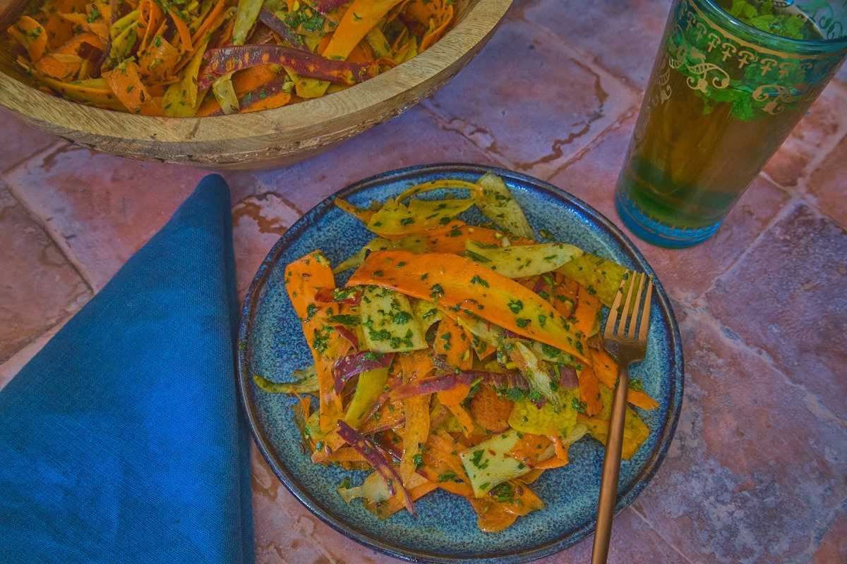 carrot ribbons in chermoula sauce on a plate with a fork, with a large wooden bowl of carrots, a glass of mint tea, and a napkin