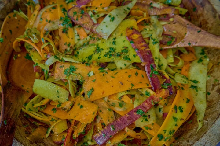 a close-up shot of carrot ribbons in chermoula sauce in a large wooden bowl with 2 wooden salad serving spoons