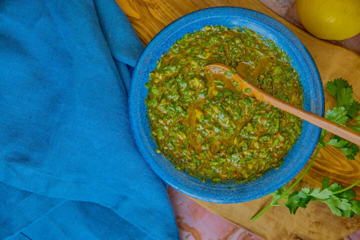 chermoula sauce in a blue bowl with a wooden spoon on a wooden board with parsley and cilantro, and a napkin and a lemon alongside
