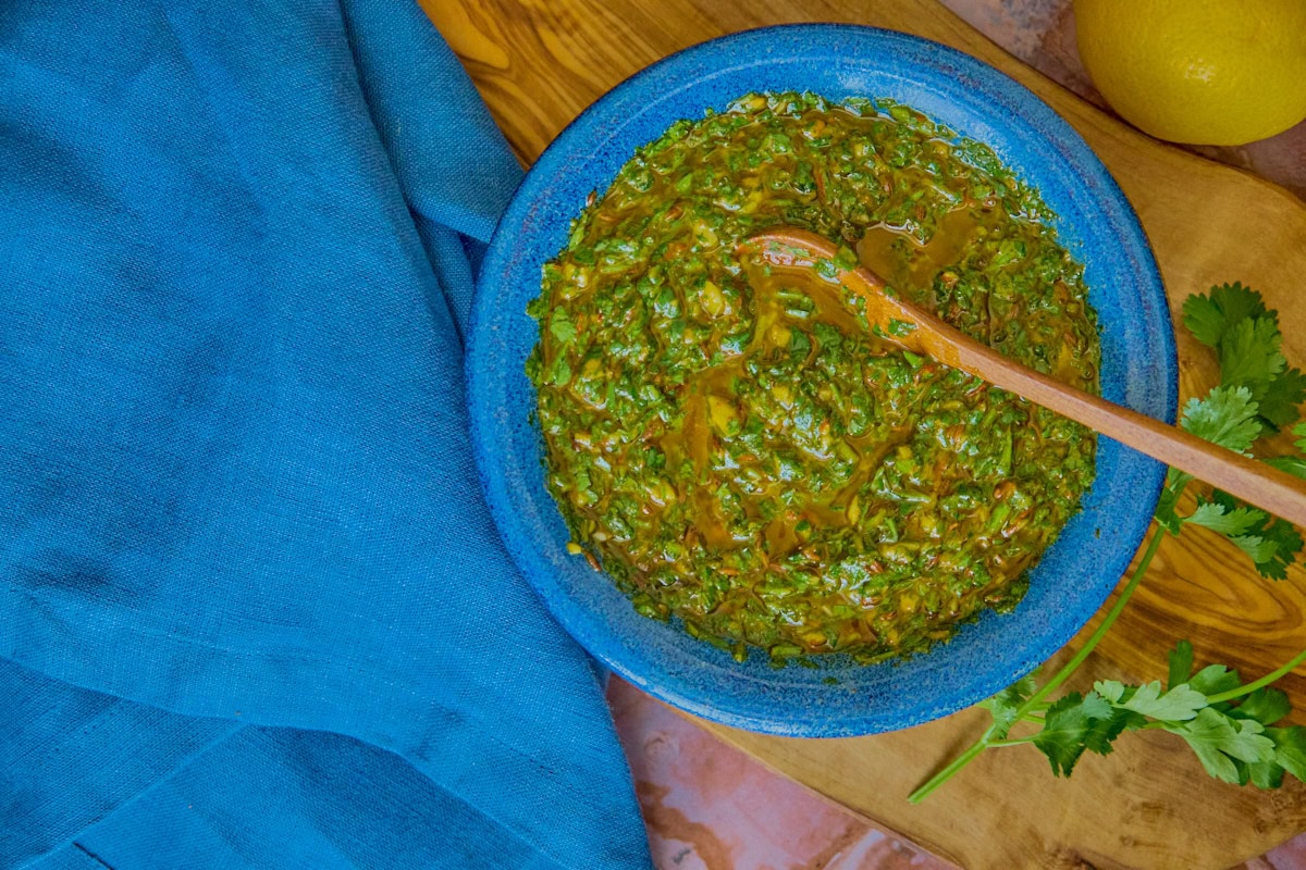 chermoula sauce in a blue bowl with a wooden spoon on a wooden board with parsley and cilantro, and a napkin and a lemon alongside