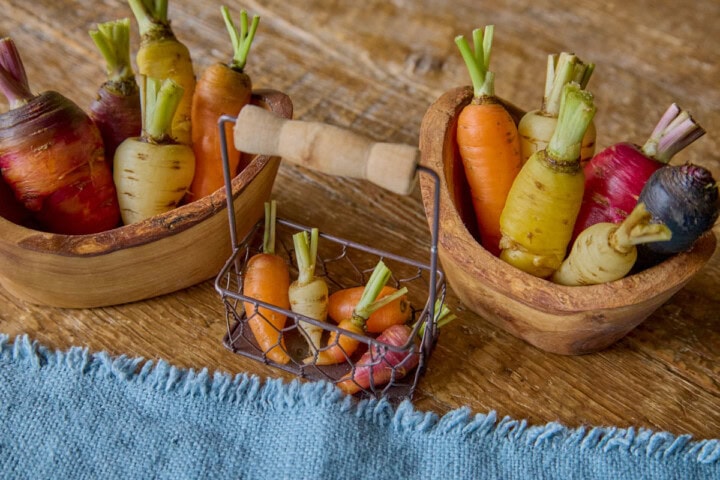 small carrots of various colors in two wooden bowls and a tiny basket, with a napkin in the foreground