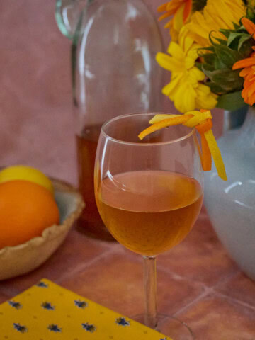 a square photo of a glass of vin d'orange with a lemon and orange strip garnish, a vase of flowers, a bowl of oranges and lemons, a napkin, and a half-full bottle