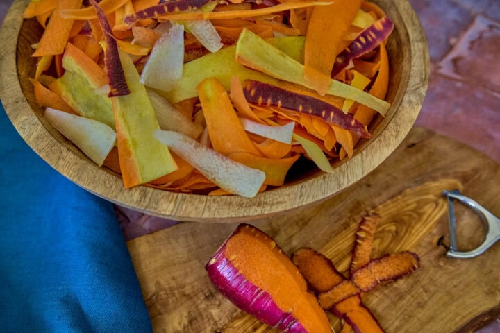 A close-up shot of carrot ribbons in a large wooden bowl next to a wooden board with a carrot, peeler, and carrot ribbons, and a napkin alongside