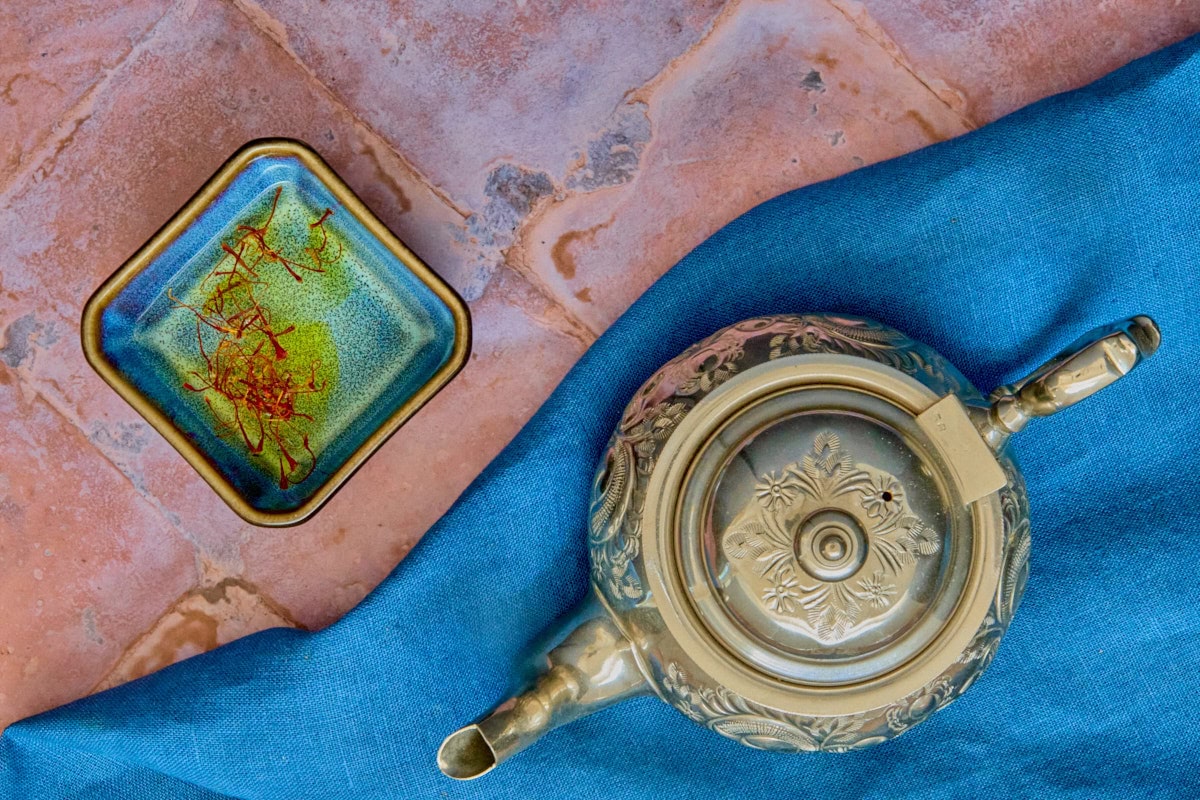saffron threads soaking in a small bowl of water, with an engraved metal teapot on a napkin in the foreground