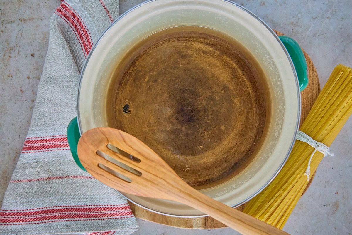 a pot of water on a wooden board with a wooden spoon on top and a tied bundle of linguini and a kitchen towel alongside