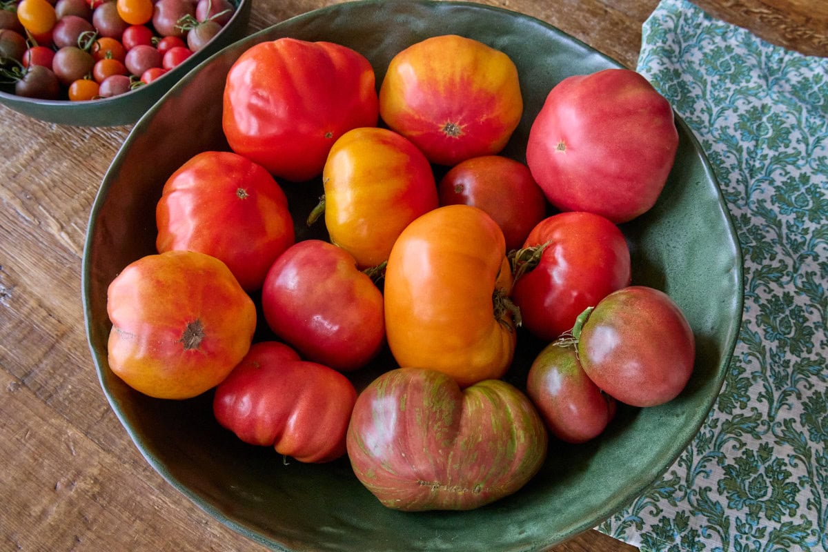 a large green bowl with large multicolored heirloom tomatoes, with a smaller green bowl of cherry tomatoes and a napkin alongside