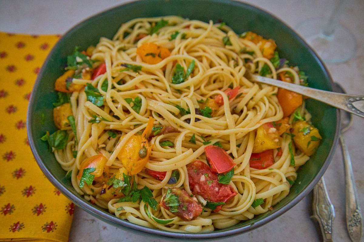 a green bowl of pasta and sauce with a fork, a napkin, knife, and spoon alongside, and a wine glass in the background