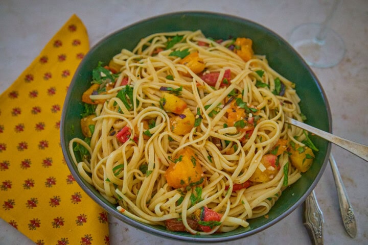 a green bowl of pasta and sauce with a fork, a napkin, knife, and spoon alongside, and a wine glass in the background