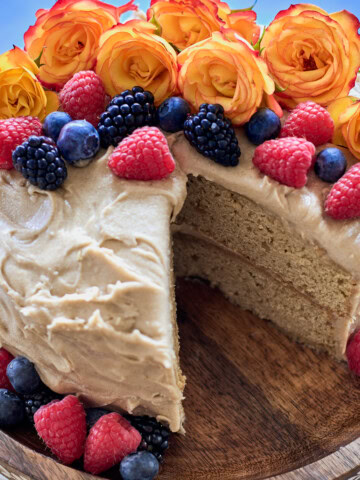 a cake on a wooden cake stand with one slice removed, and berries and roses around the cake, and a napkin in the background