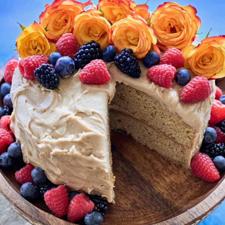 a cake on a wooden cake stand with one slice removed, and berries and roses around the cake, and a napkin in the background