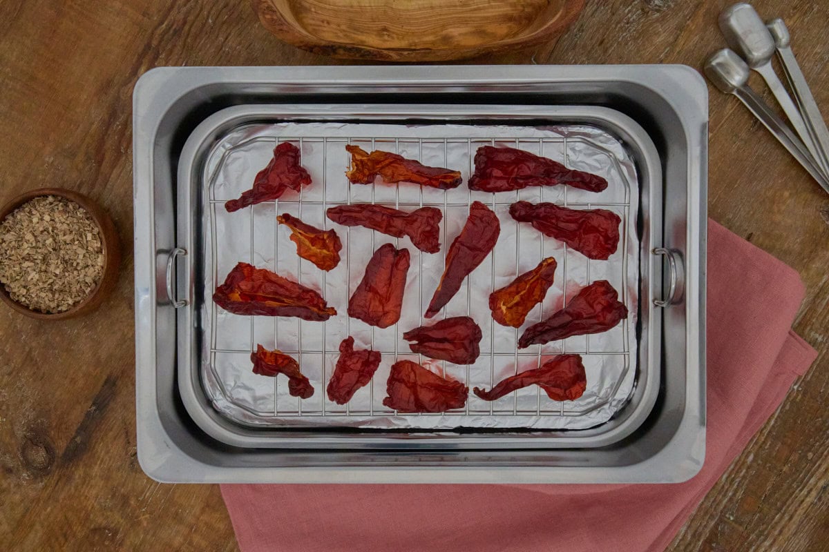 dried leutschauer peppers in a stovetop smoker with oak wood chips, measuring spoons, a wooden bowl, and a napkin alongside