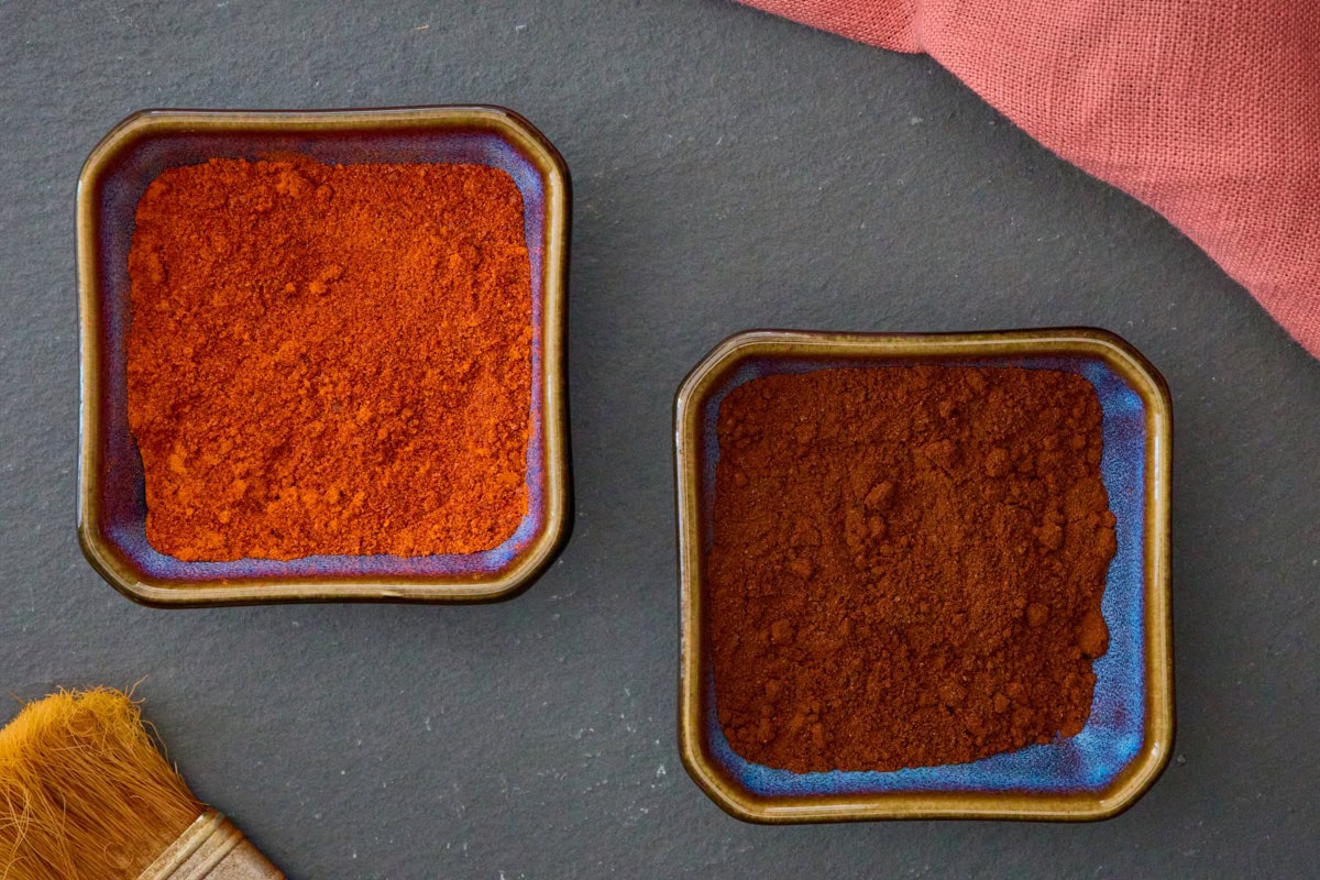 small square blue bowls of unsmoked and smoked leutschauer paprika pepper powder with a basting brush and a napkin in the background