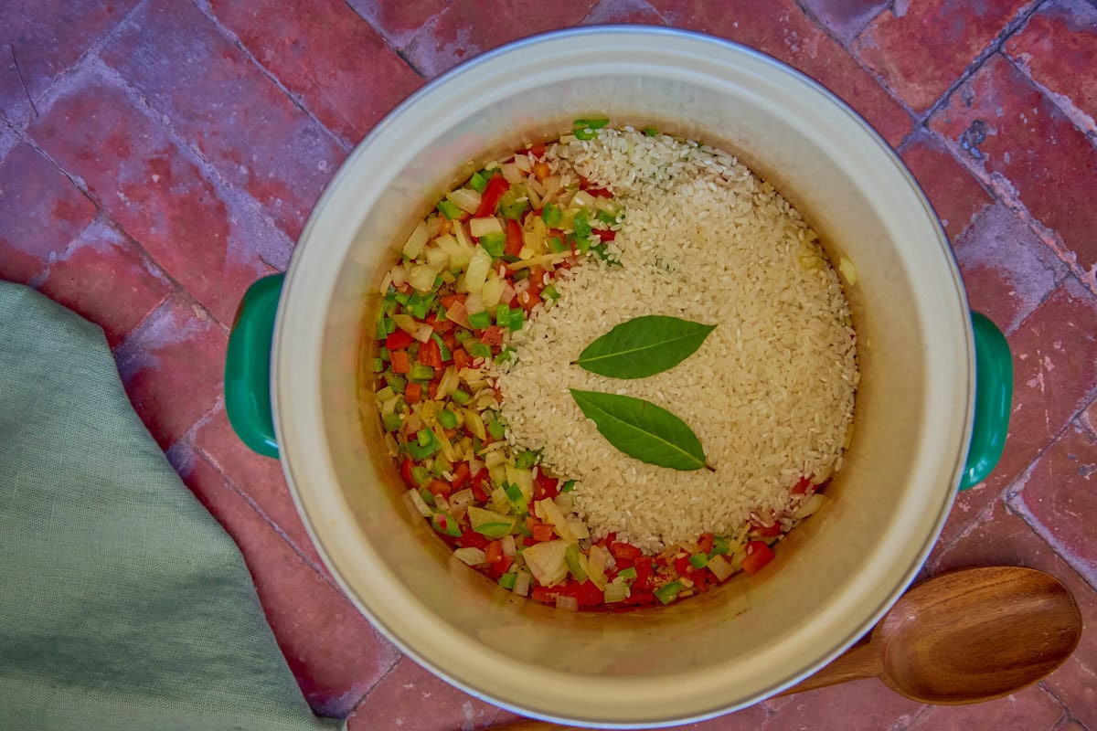 a process shot of adding rice and bay leaves to a pot full of aromatics and seasonings, with a wooden spoon and napkin in the background