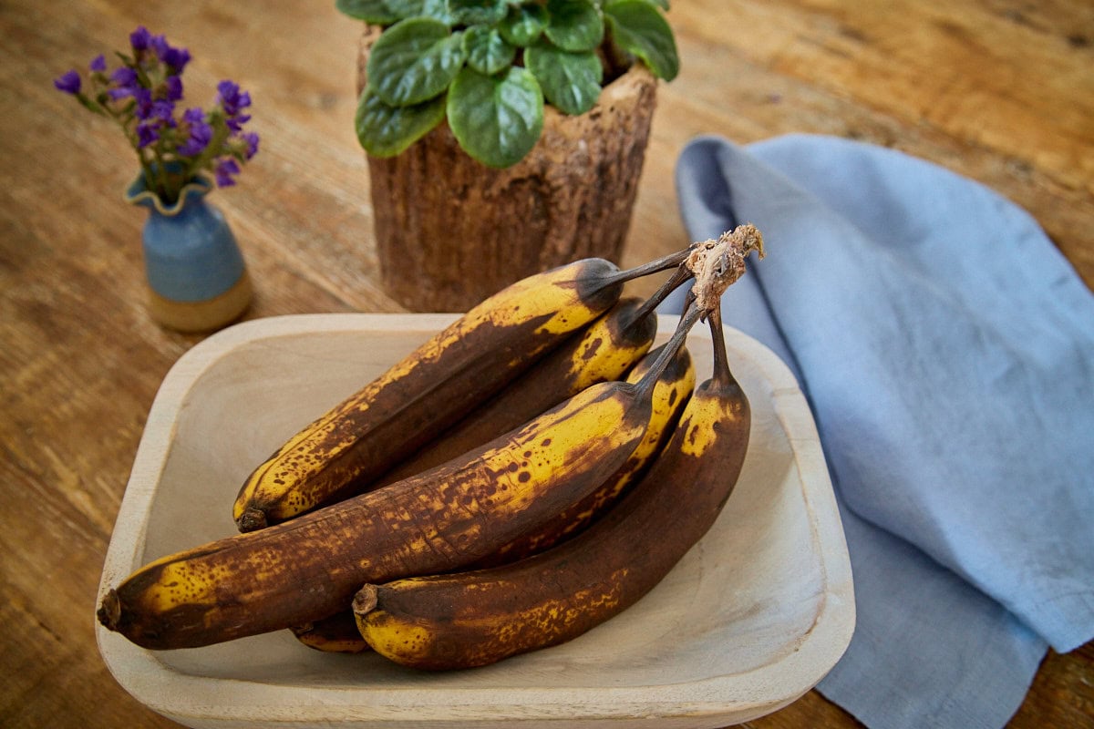 overripe bananas in a wooden bowl with a napkin, small vase of flowers, and a planter with a violet plant in the background