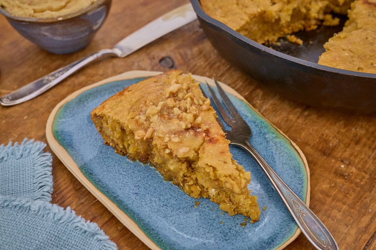a slice of banana cornbread on a plate with a fork, with a napkin, bowl of maple walnut butter, butter knife, and skillet of banana cornbread in the background