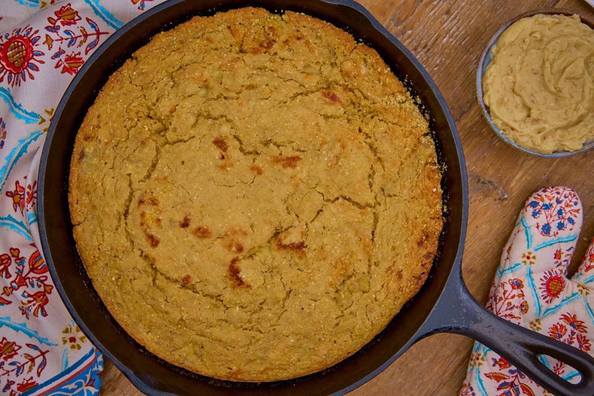 a closeup shot of banana cornbread in a skillet, a bowl of maple walnut butter, an oven mitt, and a kitchen towel in the background