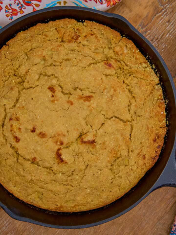baked banana cornbread in a cast iron skillet, with a bowl of maple walnut butter, an oven mitt, and a kitchen table alongside