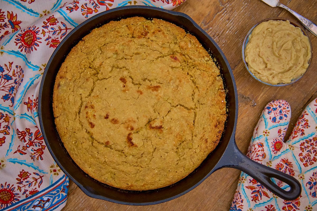 banana cornbread in a skillet with a bowl of maple walnut butter with a butter knife, an oven mitt, and a kitchen towel in the background