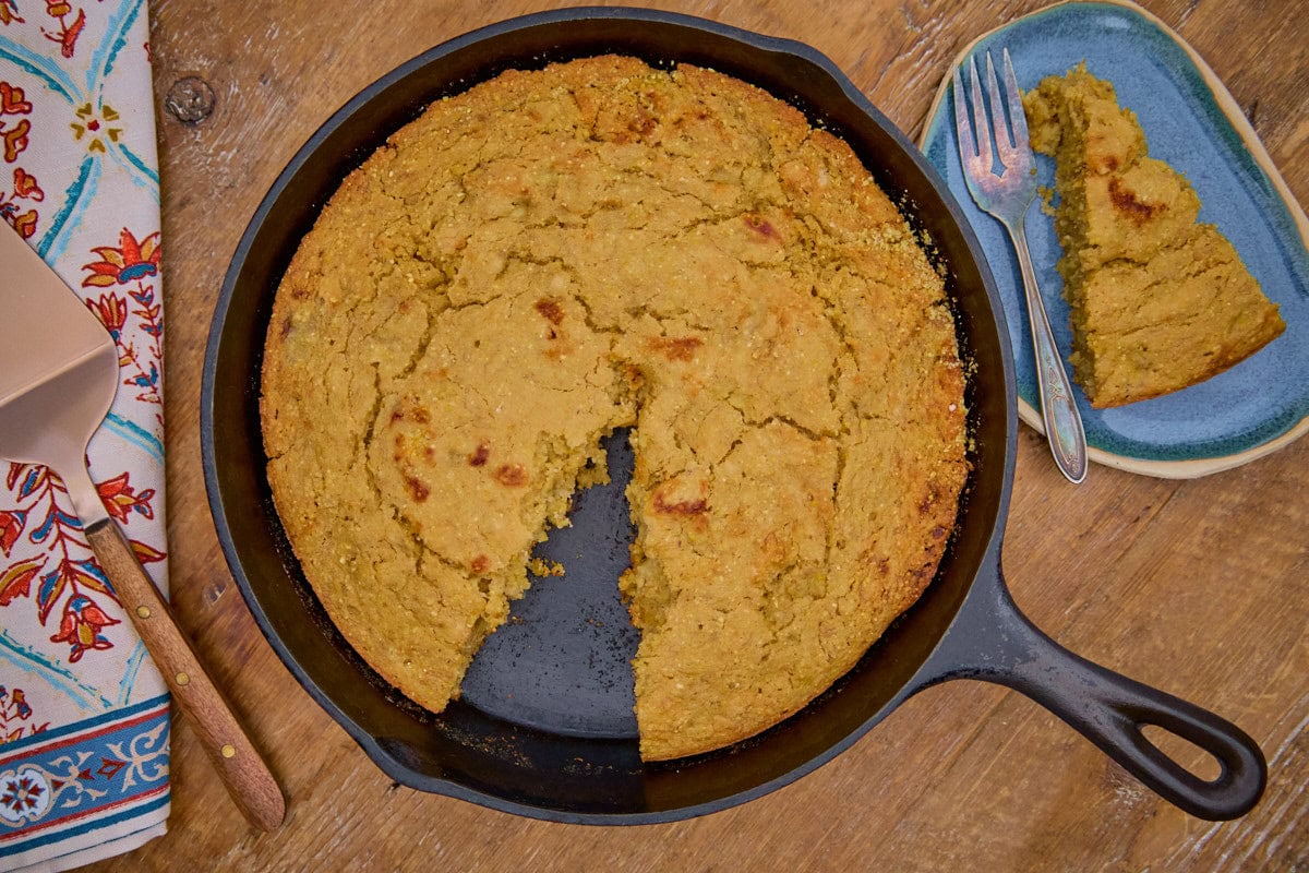 banana cornbread in a skillet with a slice removed, with the slice on a plate with a fork, and a cake server and kitchen towel on the side