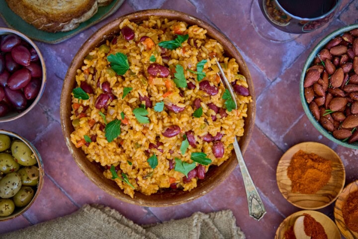 a bowl of beans and rice with a piece of bread, olives, almonds, paprika, a glass of wine, a napkin, and a fork in the background