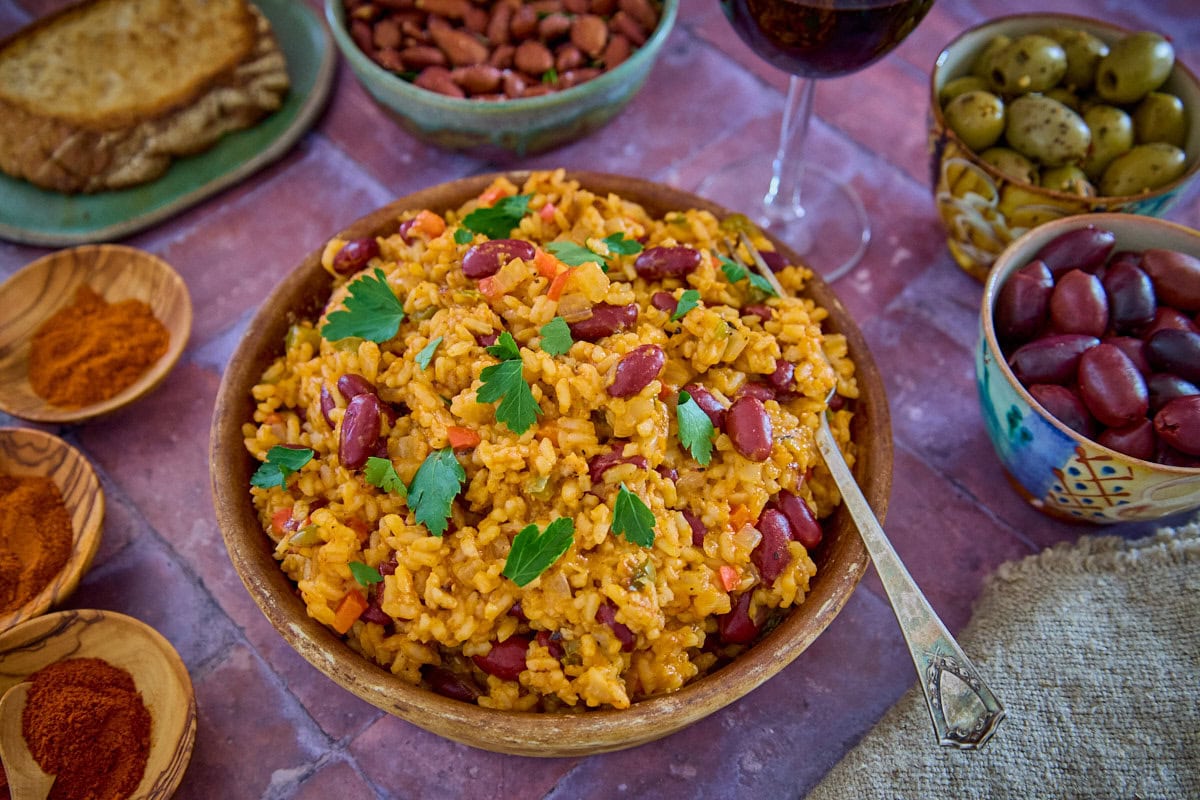 a bowl of beans and rice with a piece of bread, olives, almonds, paprika, a glass of wine, a napkin, and a fork in the background