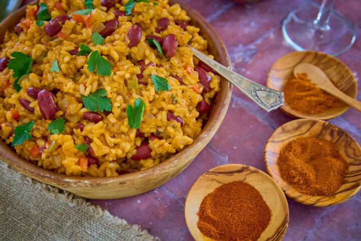 a bowl of beans and rice with three types of pepper powders, a glass of wine, and a napkin in the background