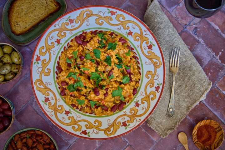 a bowl of beans and rice with a piece of bread, olives, almonds, paprika, a glass of wine, a napkin, and a fork in the background