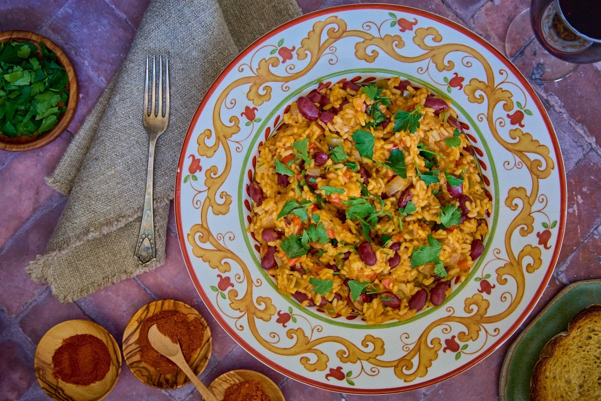 a bowl of beans and rice with a piece of bread, parsley, paprika, a glass of wine, a napkin, and a fork in the background