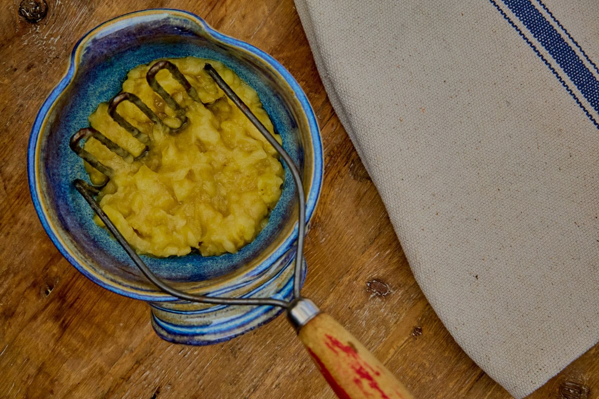 mashed bananas in a small mixing bowl with a potato masher, and a kitchen towel in the background