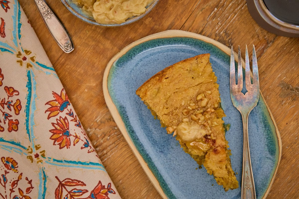 a slice of banana cornbread on a plate with a fork, with a kitchen towel, bowl of maple walnut butter, butter knife, and cup of coffee in the background