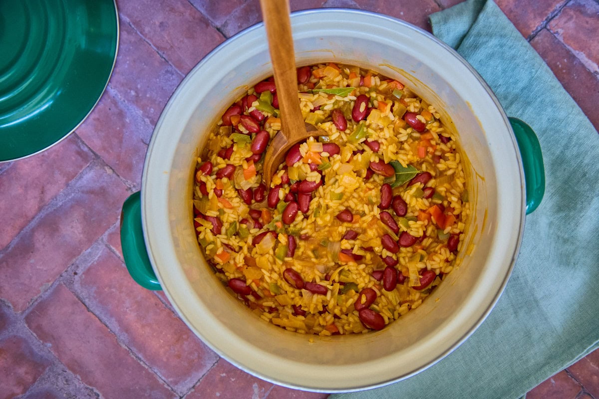 a process shot of adding beans to a pot with rice, liquids, aromatics and seasonings, with a wooden spoon and napkin in the background