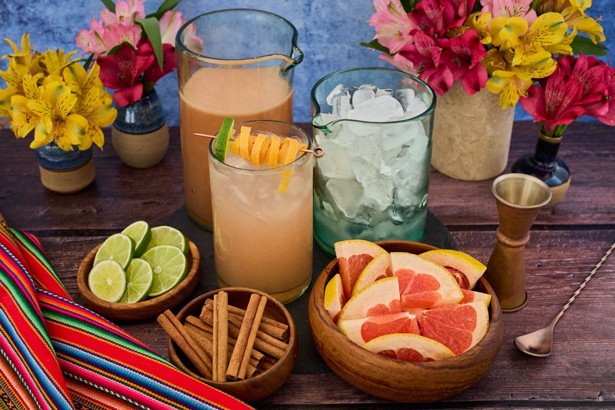 a chirulin spritz cocktail in a glass, with vases of flowers, a mixing glass full of additional cocktail, a mixing glass of ice, a jigger, mixing spoon, bowls of grapefruit slices, cinnamon sticks, and lime slices, and a piece of fabric