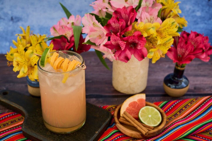 a chirulin spritz cocktail in a glass on a wooden board, with vases of flowers, a bowl with a grapefruit slice, cinnamon sticks, and lime slice, and a piece of fabric