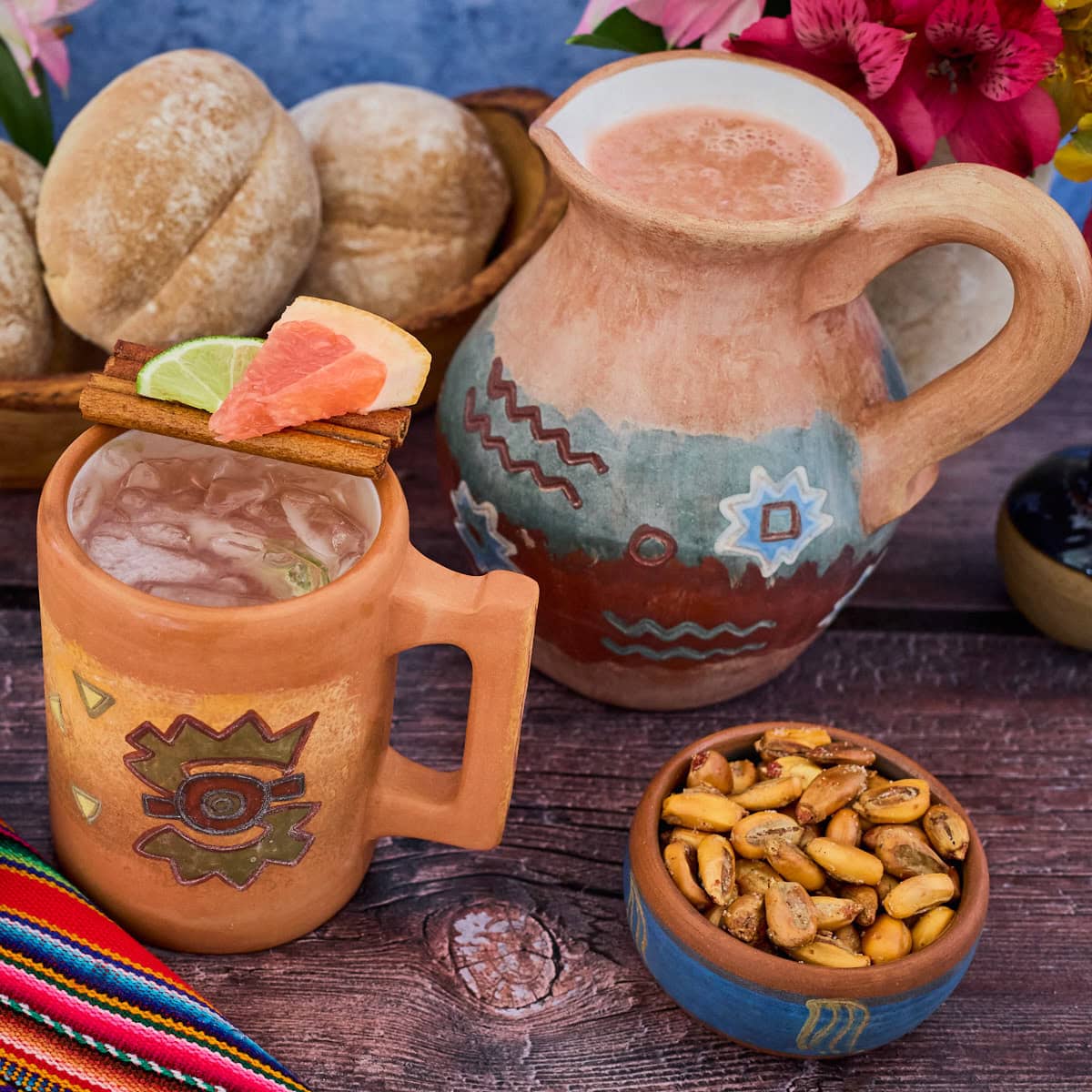 a Peruvian clay mug and pitcher of chirulin spritz cocktail with bread rolls, vases of flowers, toasted corn in a bowl, and a piece of fabric around them