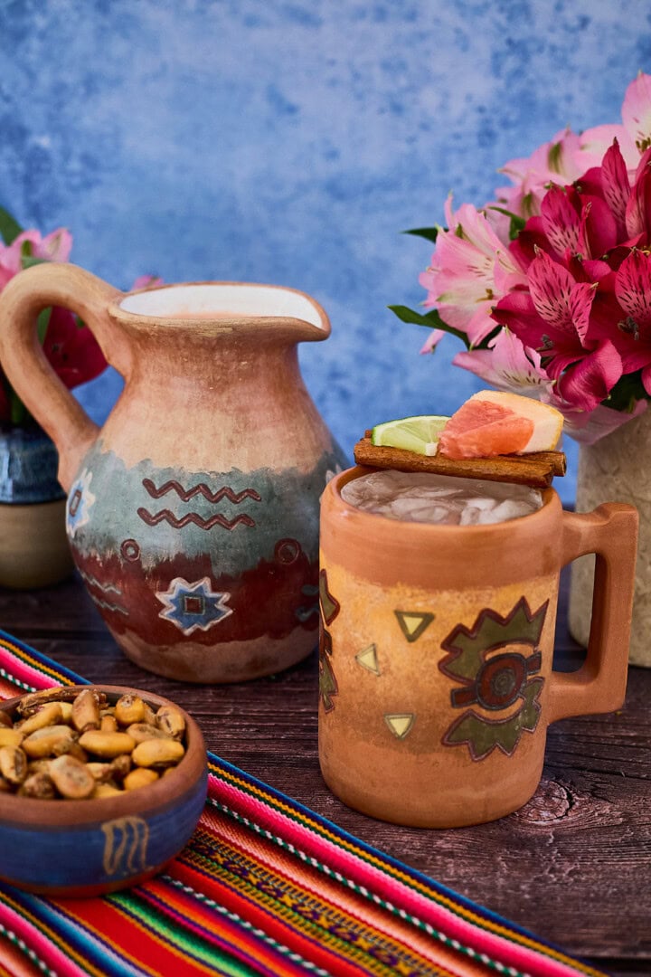 a Peruvian clay mug and pitcher of chirulin spritz cocktail with vases of flowers, toasted corn, and a piece of fabric around them