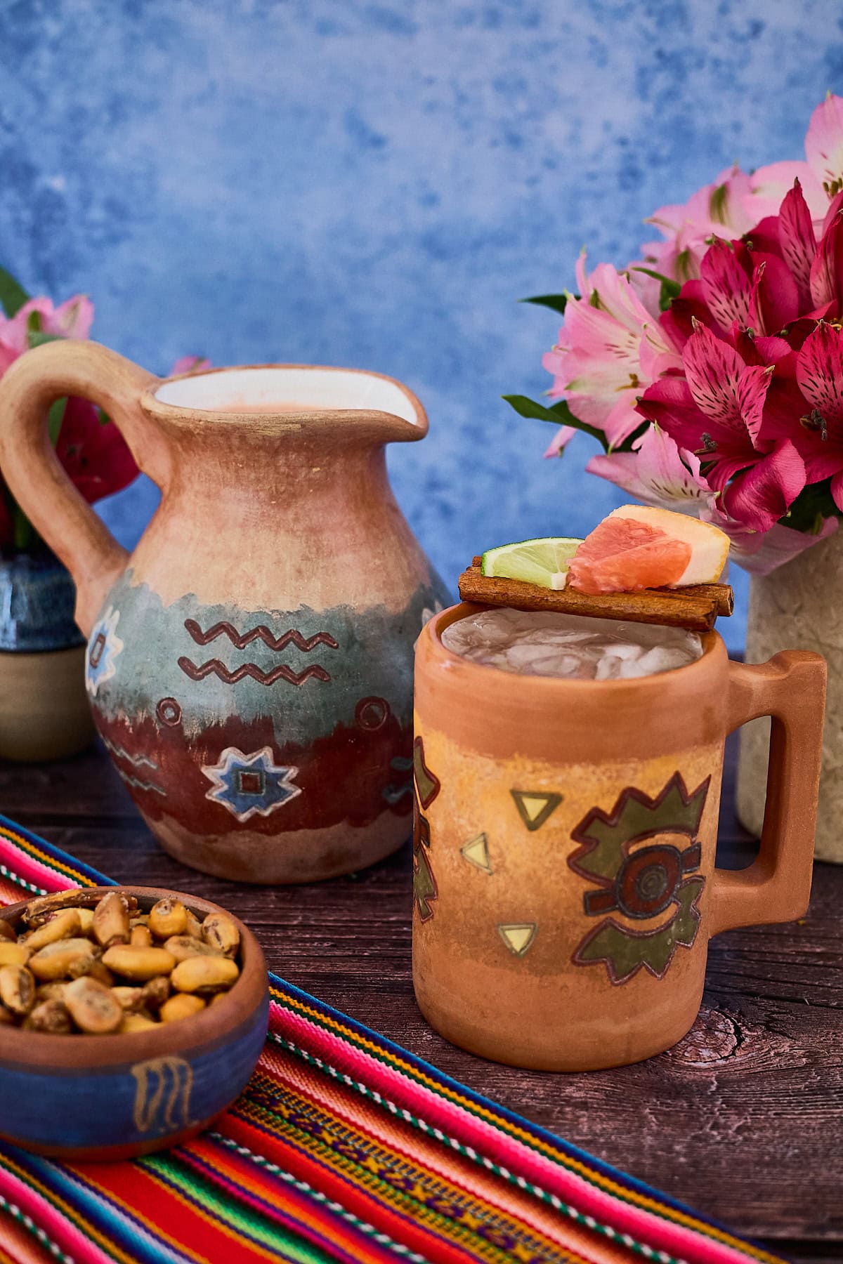 a Peruvian clay mug and pitcher of chirulin spritz cocktail with vases of flowers, toasted corn, and a piece of fabric around them