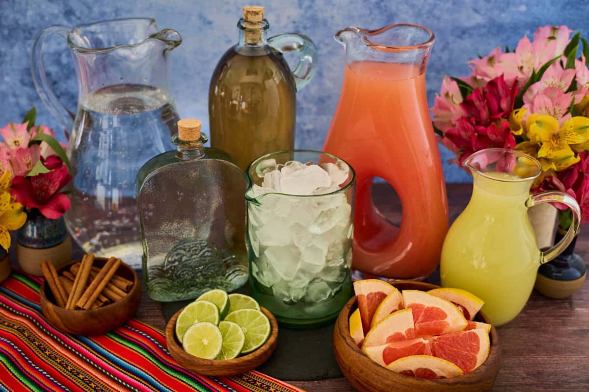 a bottle of pisco, pitcher of sparkling water, pitcher of grapefruit juice, pitcher of lime juice, vases of flowers, a mixing glass of ice, bowls of grapefruit slices, cinnamon sticks, and lime slices, and a piece of fabric