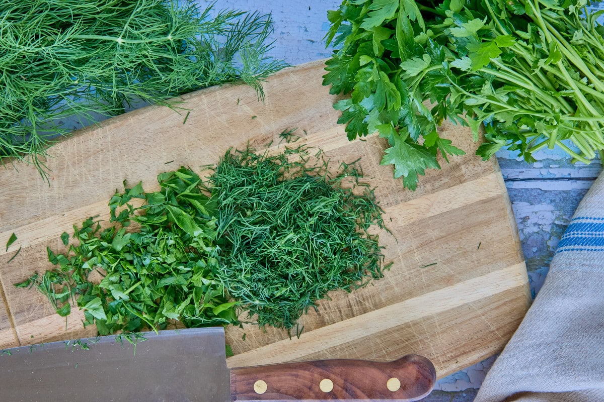 chopped dill and parsley on a cutting board with a knife, kitchen towel, and bundles of parsley and dill