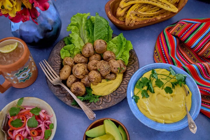 huancaína sauce in a bowl, surrounded by a piece of fabric, toasted plantain chips, a plate of lettuce, roasted baby potatoes with huancaína sauce, avocado slices, a mug of chirulín cocktail, a vase of flowers, and pickled onions and chilis