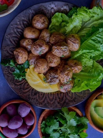 huancaína sauce in a bowl, surrounded by a piece of fabric, pickled onions and chilis, a plate of lettuce and roasted baby potatoes, olives, vase of flowers, huancaína sauce in a bowl, a piece of fabric, lime wedges, avocado slices, olives, vase of flowers, chirulín cocktail, and cilantro