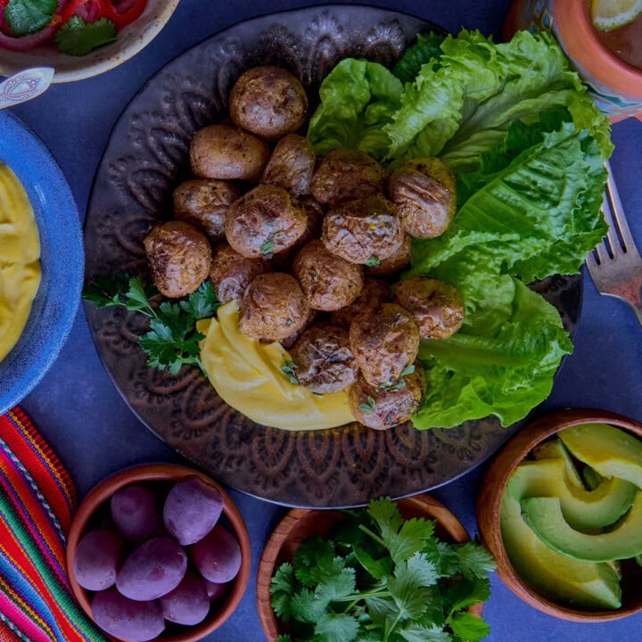 huancaína sauce in a bowl, surrounded by a piece of fabric, pickled onions and chilis, a plate of lettuce and roasted baby potatoes, olives, vase of flowers, huancaína sauce in a bowl, a piece of fabric, lime wedges, avocado slices, olives, vase of flowers, chirulín cocktail, and cilantro