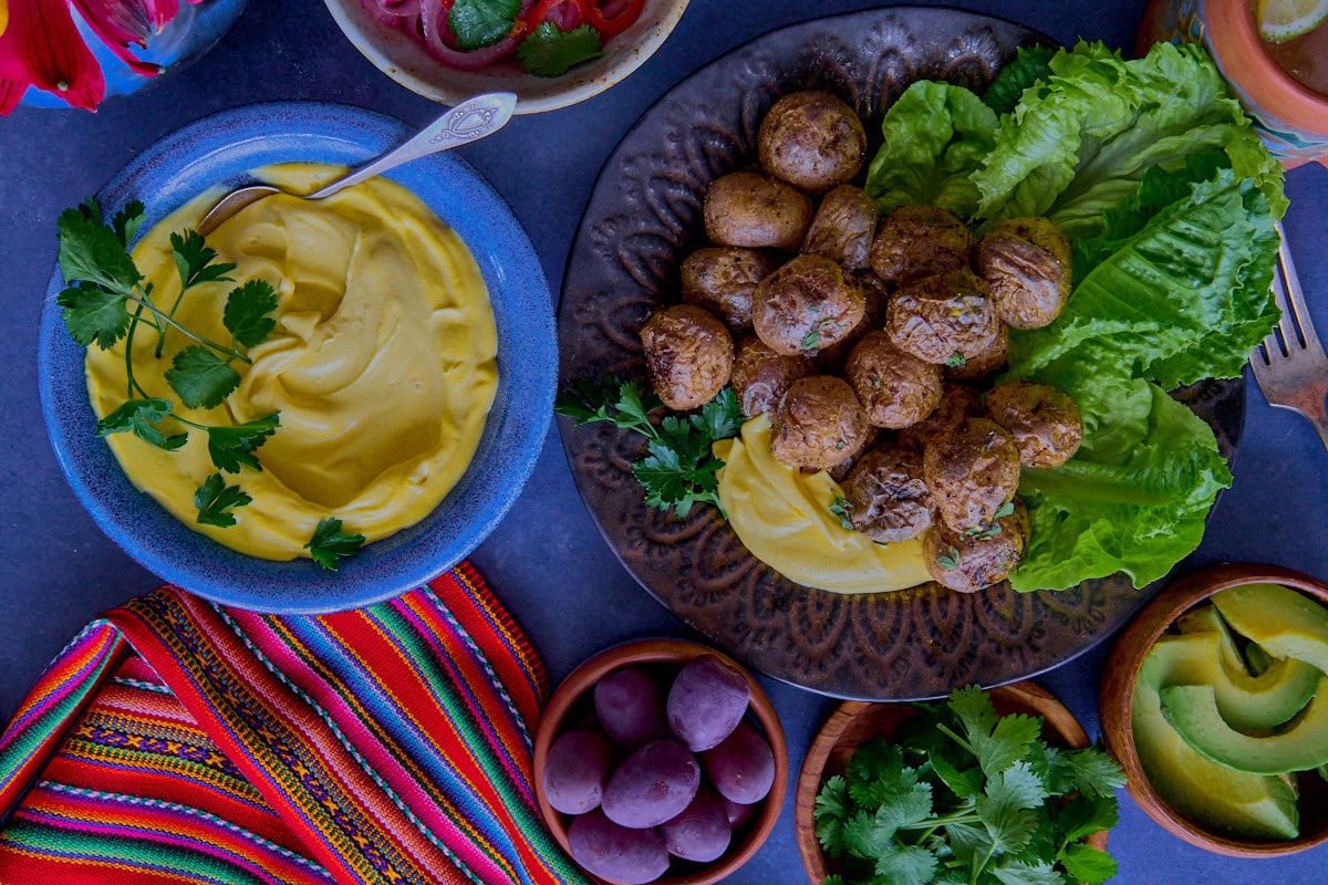 huancaína sauce in a bowl, surrounded by a piece of fabric, pickled onions and chilis, a plate of lettuce and roasted baby potatoes, olives, vase of flowers, avocado slices, olives, vase of flowers, chirulín cocktail, and cilantro