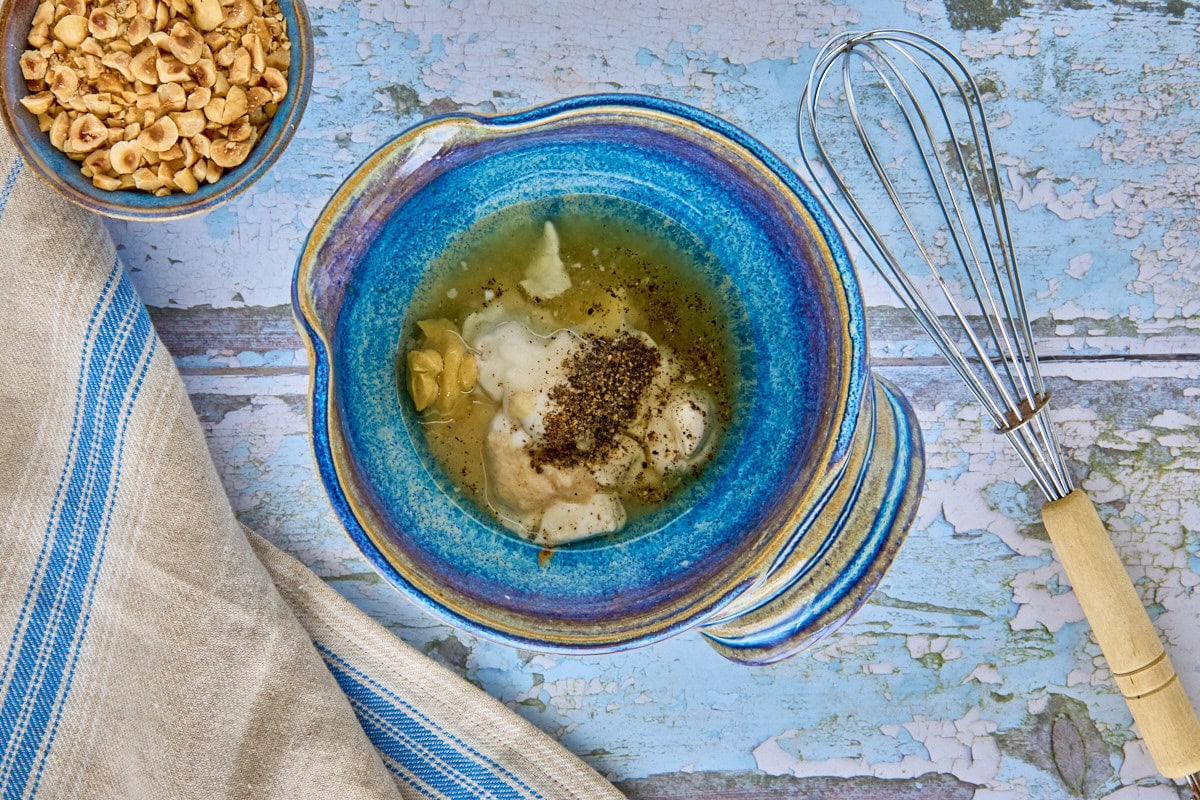 a bowl of dressing ingredients, bowl of toasted hazelnuts, kitchen towel, and whisk